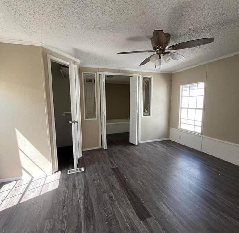 Empty room with light brown walls, dark wood flooring, a ceiling fan, and a large window on the right. Two open doorways reveal a closet and bathroom. Bright, inviting ambiance.