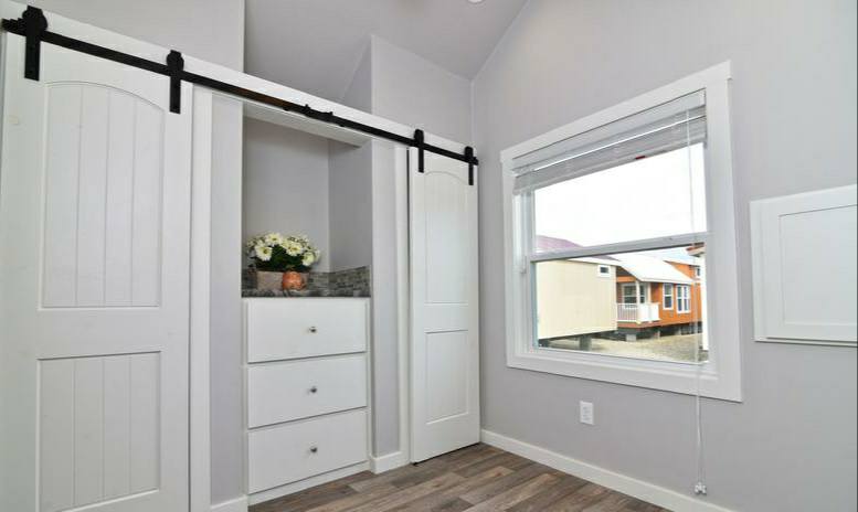 Bright room with white sliding barn doors revealing shelves, white drawers, and a vase of flowers. A large window shows a view of neighboring houses.