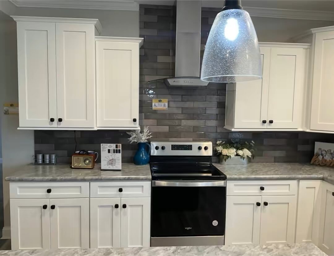 A modern kitchen with white cabinets, a glossy black oven, gray tile backsplash, and marble countertops. There's a bright pendant light, flowers, and decor creating a welcoming atmosphere.