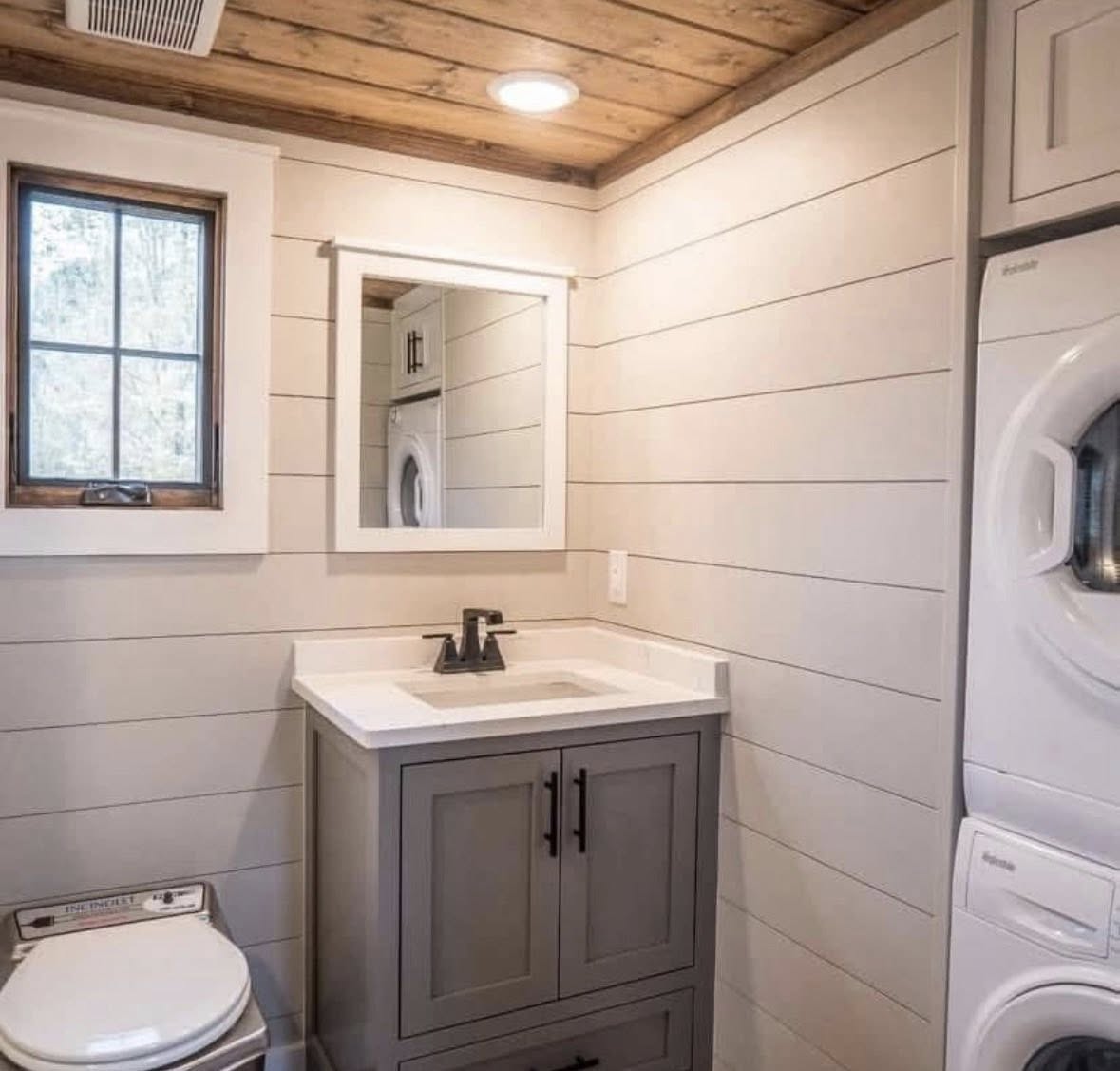 A compact bathroom with a wooden ceiling and light-colored shiplap walls, featuring a small window, a mirror above a sink, and a stacked washer and dryer.