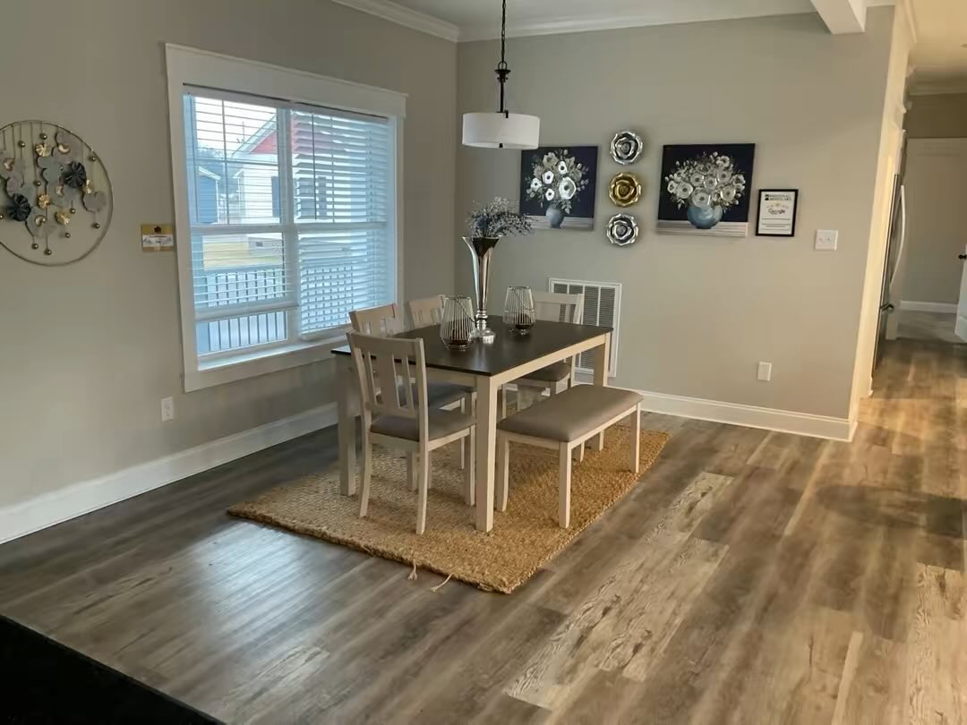 A cozy dining area with a dark wooden table, four chairs, and a bench on a woven rug. Art and decorative plates adorn the light gray walls. Natural light filters in through a window, creating a warm and inviting atmosphere.