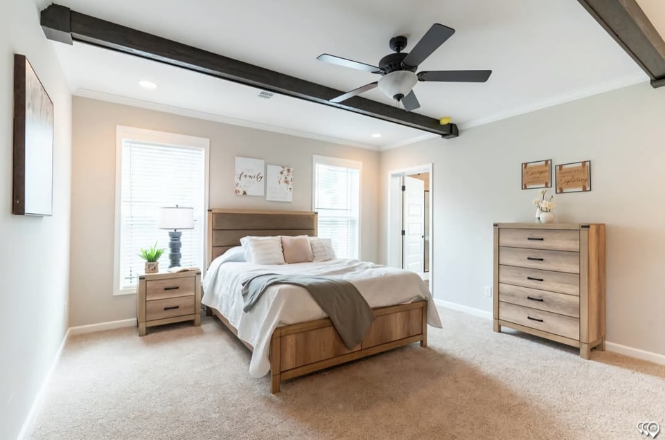 A cozy bedroom with neutral tones features a wooden bed, matching nightstand, and dresser. Large windows brighten the room, conveying a serene ambiance.