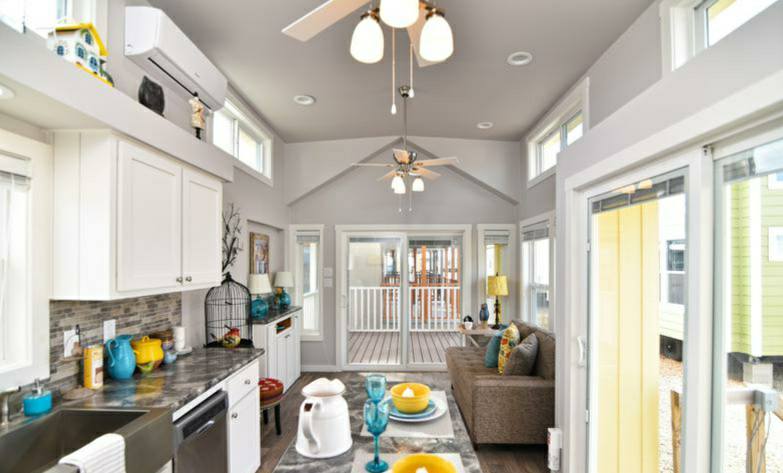 Bright, modern tiny home interior with white cabinets, colorful accents, a gray sofa, and large windows. Airy, cozy, and inviting atmosphere.