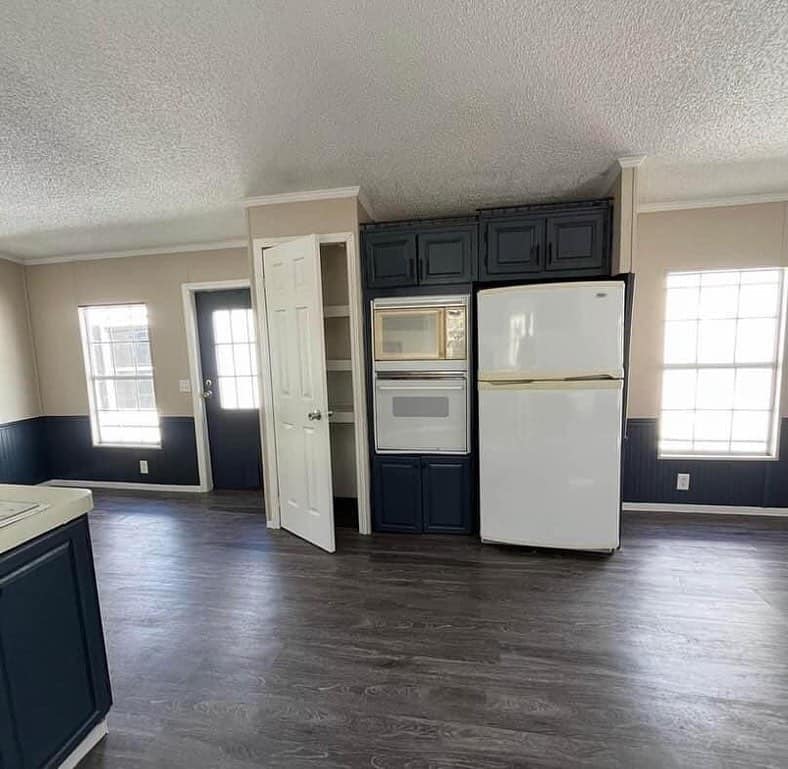 A modern kitchen space with dark wood flooring, a white fridge, an oven, and navy cabinets. Two large windows and a door allow natural light.