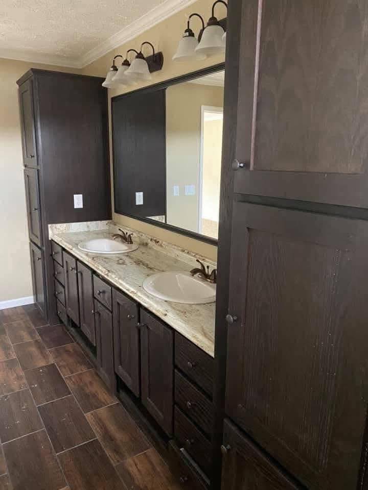 Dark wood bathroom vanity with two sinks and a large mirror. Overhead, three-light fixture illuminates the space. Brown tiled floor adds warmth.