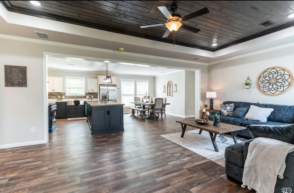 Open-concept living room and kitchen with dark hardwood floors, blue-gray sectional, wooden coffee table, elegant decor, and ceiling fan. Bright, inviting atmosphere.