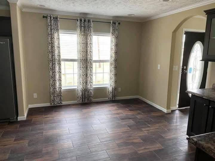 Empty room with beige walls and dark wooden floors. Two windows with patterned curtains brighten the space, next to a decorative oval door. Calm ambiance.