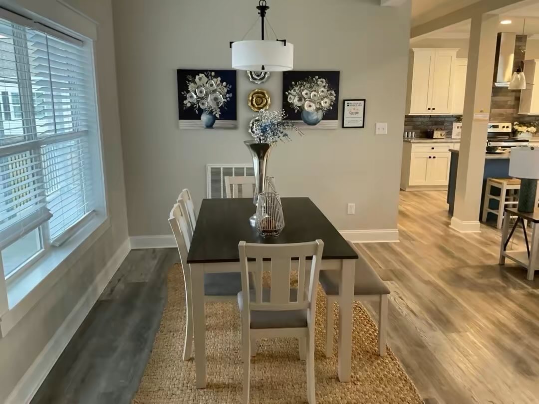 Bright dining room with a modern table set, white chairs, and vase centerpiece on a woven rug. Art on walls and soft lighting create a cozy atmosphere.