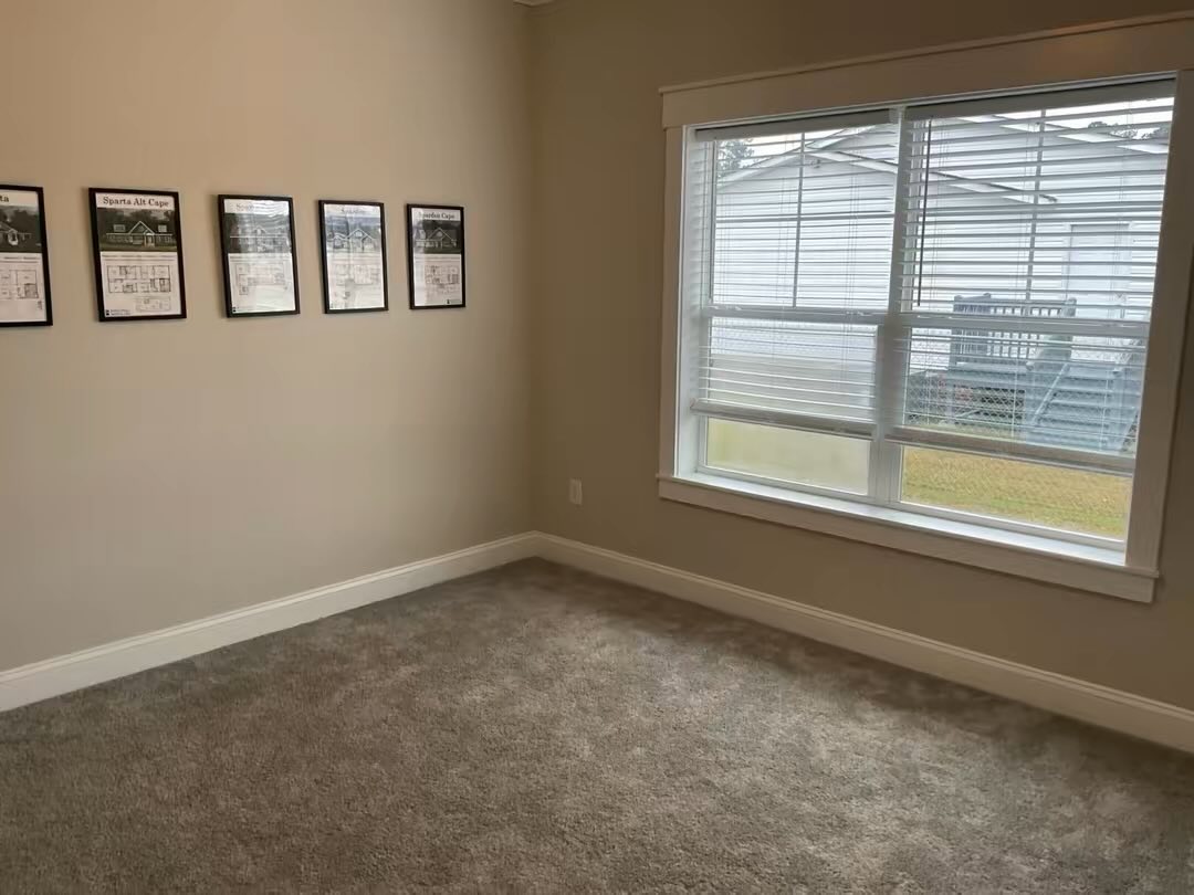 Empty room with beige walls, carpeted floor, and a large window on the right. Five framed posters hang on the left wall, conveying a minimalist tone.