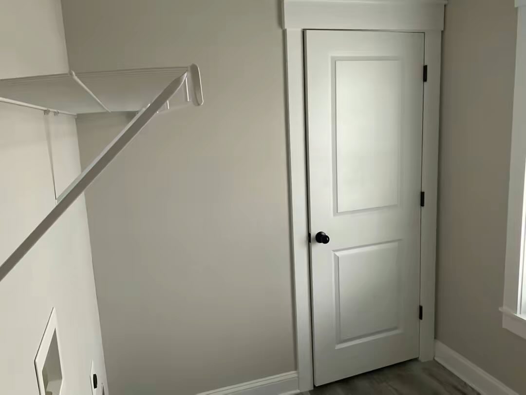 Empty laundry room with light gray walls, white door with dark handle, and a white wire shelf. Bright and clean with natural light from a window.