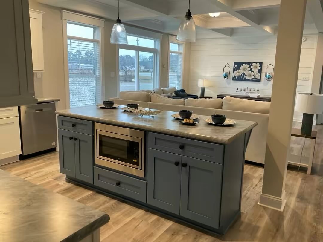 Modern open-concept kitchen and living area with a blue island featuring a microwave. Beige sofas and floral artwork add a cozy touch. Warm, inviting tone.