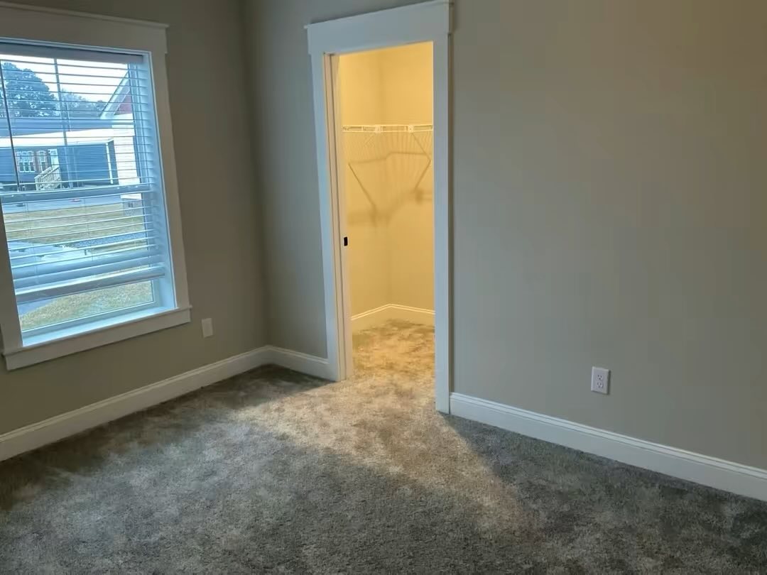 Empty room with gray carpet, white walls, and a window showing an outdoor view. Open closet door with hangers, evoking a neutral, clean tone.
