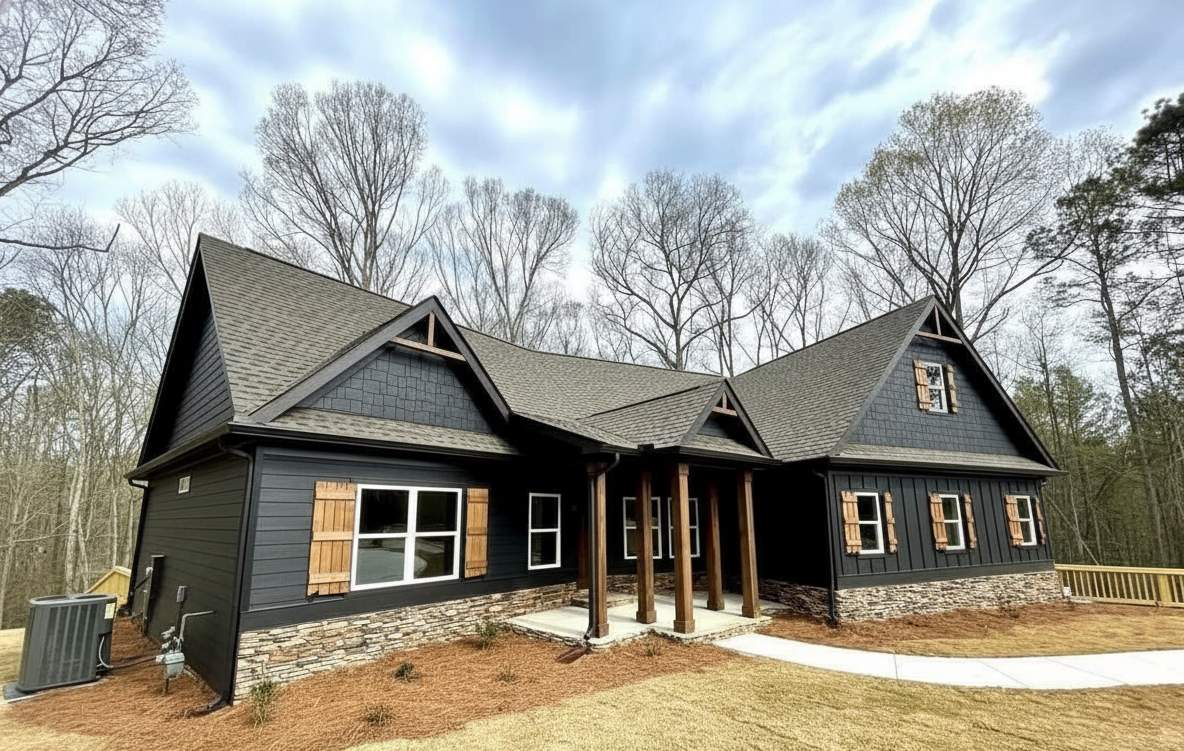 A modern, dark-colored house with stone accents and wooden shutters is set against a backdrop of bare trees under a cloudy sky, conveying a serene, rustic vibe.