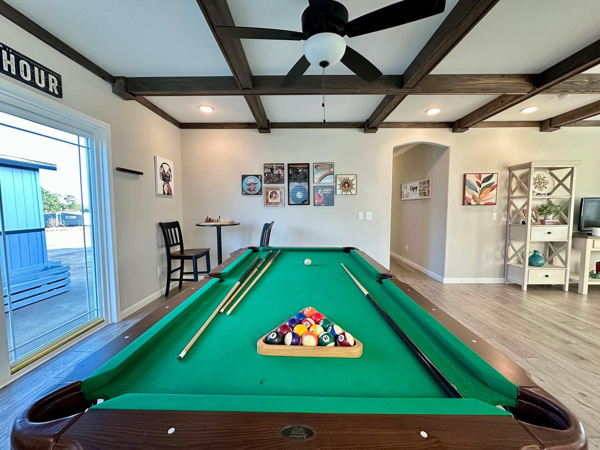 A cozy game room with a green felt pool table in the center, billiard balls racked, and cue sticks resting on the table. Wall art and a shelf add a relaxed, inviting feel.