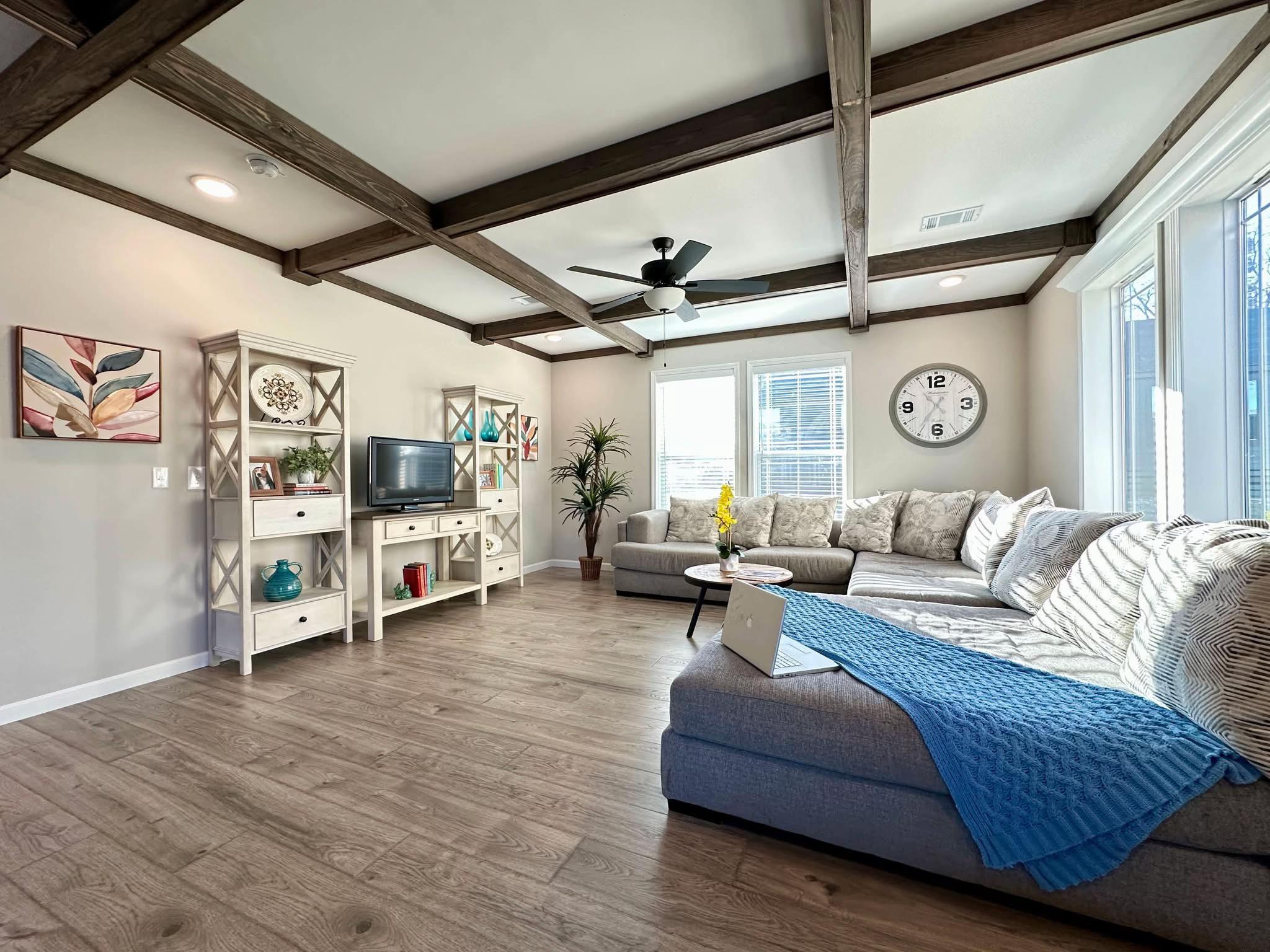 Spacious living room with wooden beams, gray sectional, blue throw, wall clock, and plant. Shelves hold decor. Bright windows, cozy ambiance.