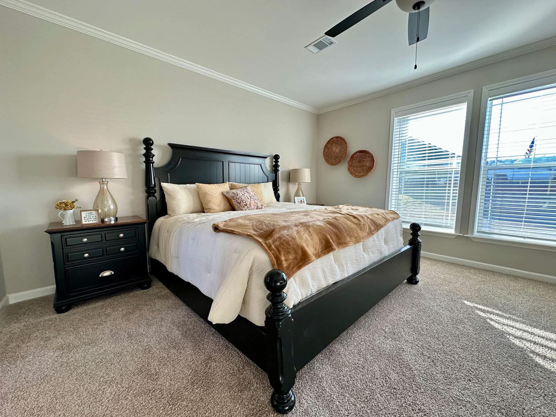 Spacious bedroom with a large black bed, fluffy pillows, and a brown throw blanket. Light filters through two large windows, creating a cozy, serene atmosphere.