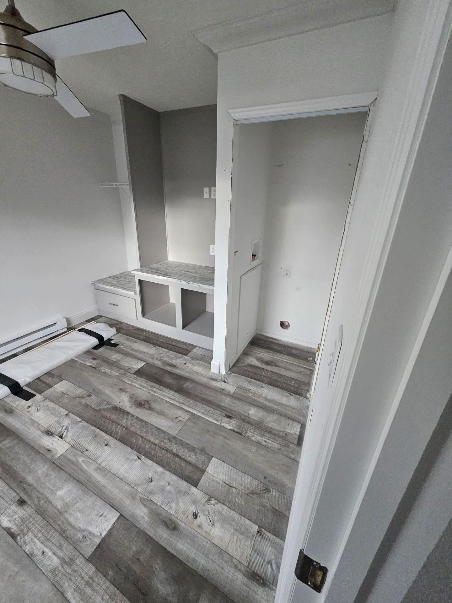 A small room with gray wood flooring, partially unrolled rug, and a built-in desk with drawers. A white ceiling fan and soft gray walls create a calm atmosphere.