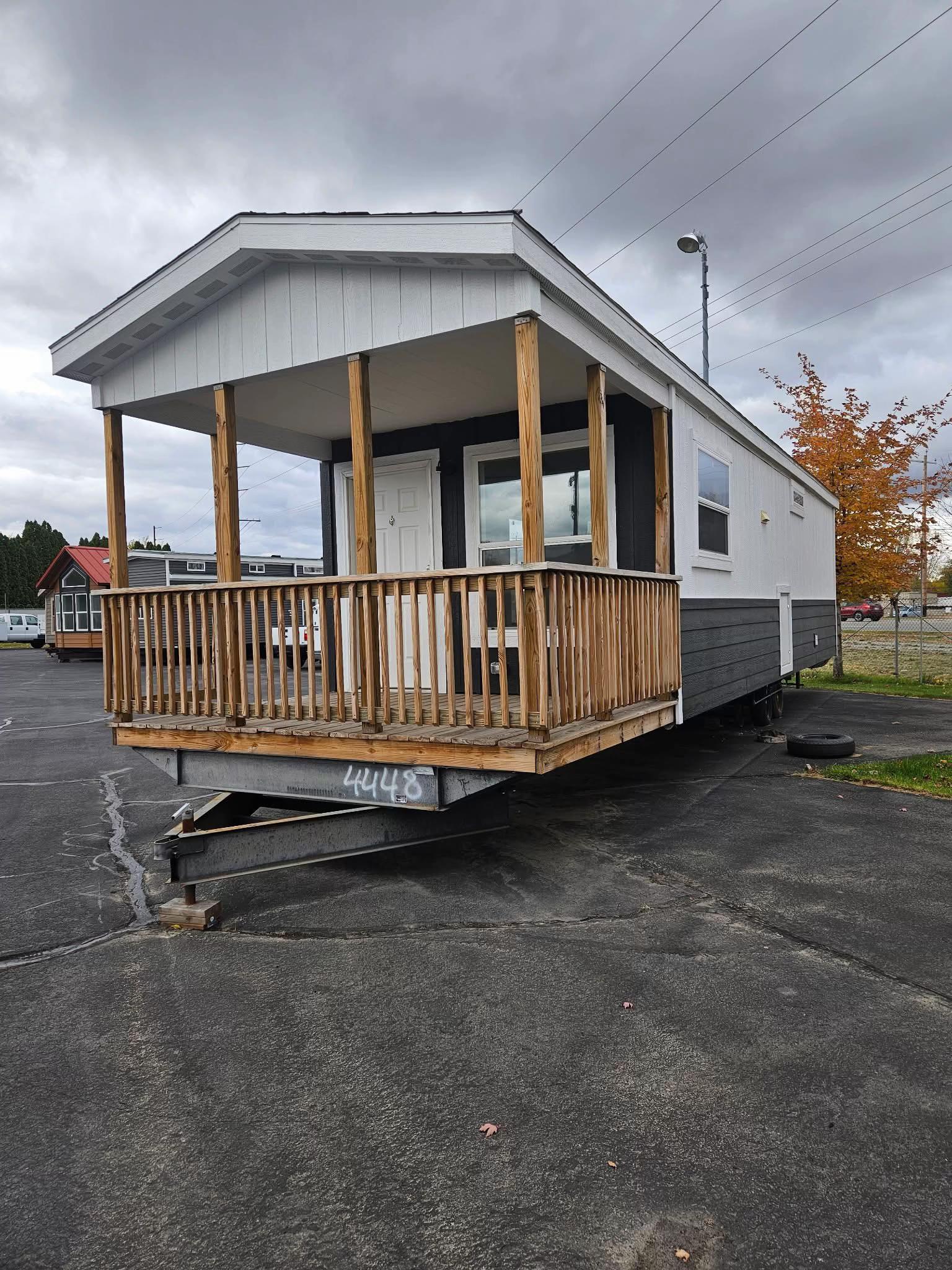 A compact mobile home parked on a paved lot. It features a small wooden porch with a railing, a white door, and large windows. Cloudy sky above.