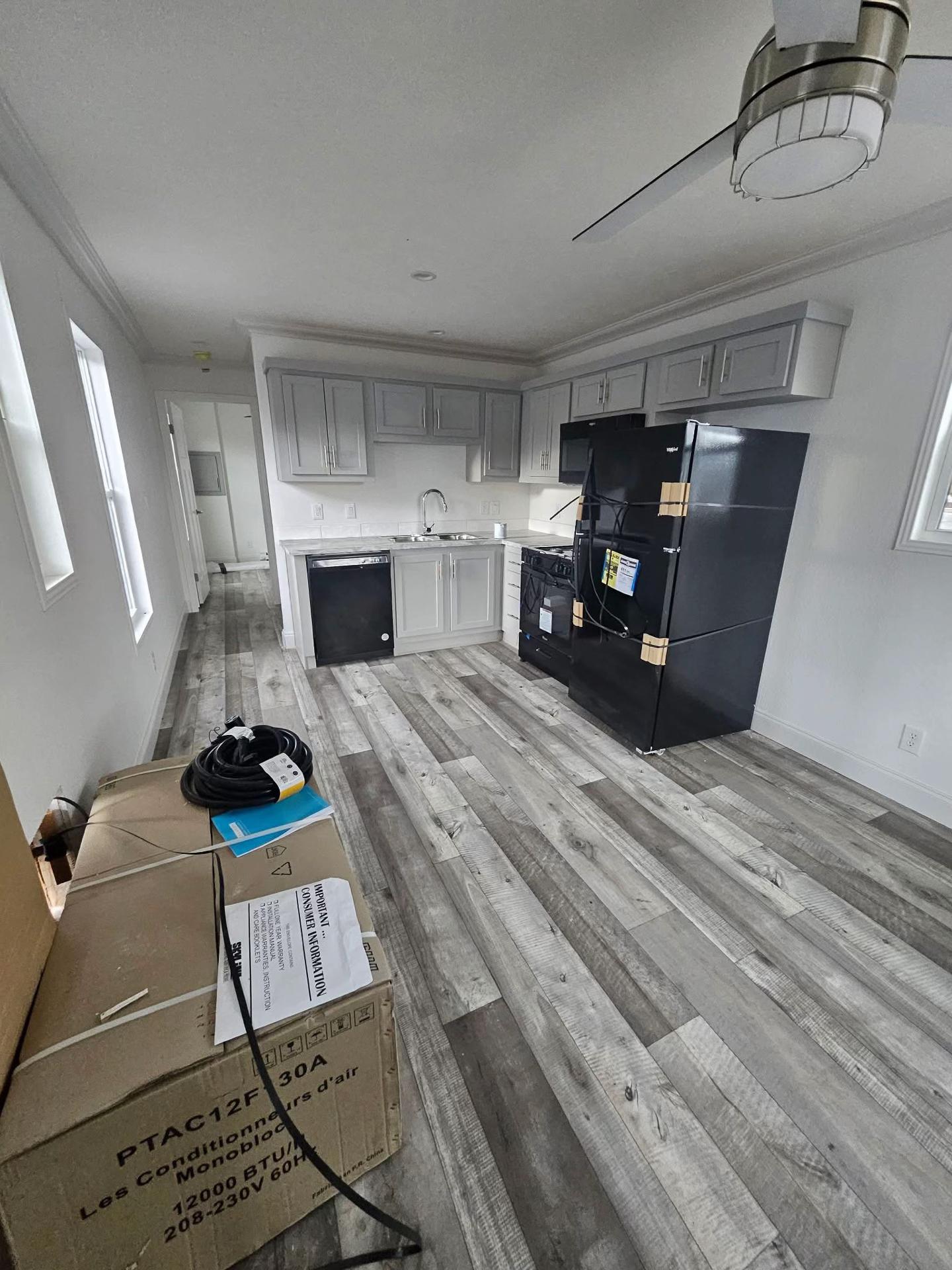 Small kitchen with gray cabinets, black appliances, and wood-patterned vinyl flooring. A ceiling fan is visible. Packaging boxes are in the foreground.