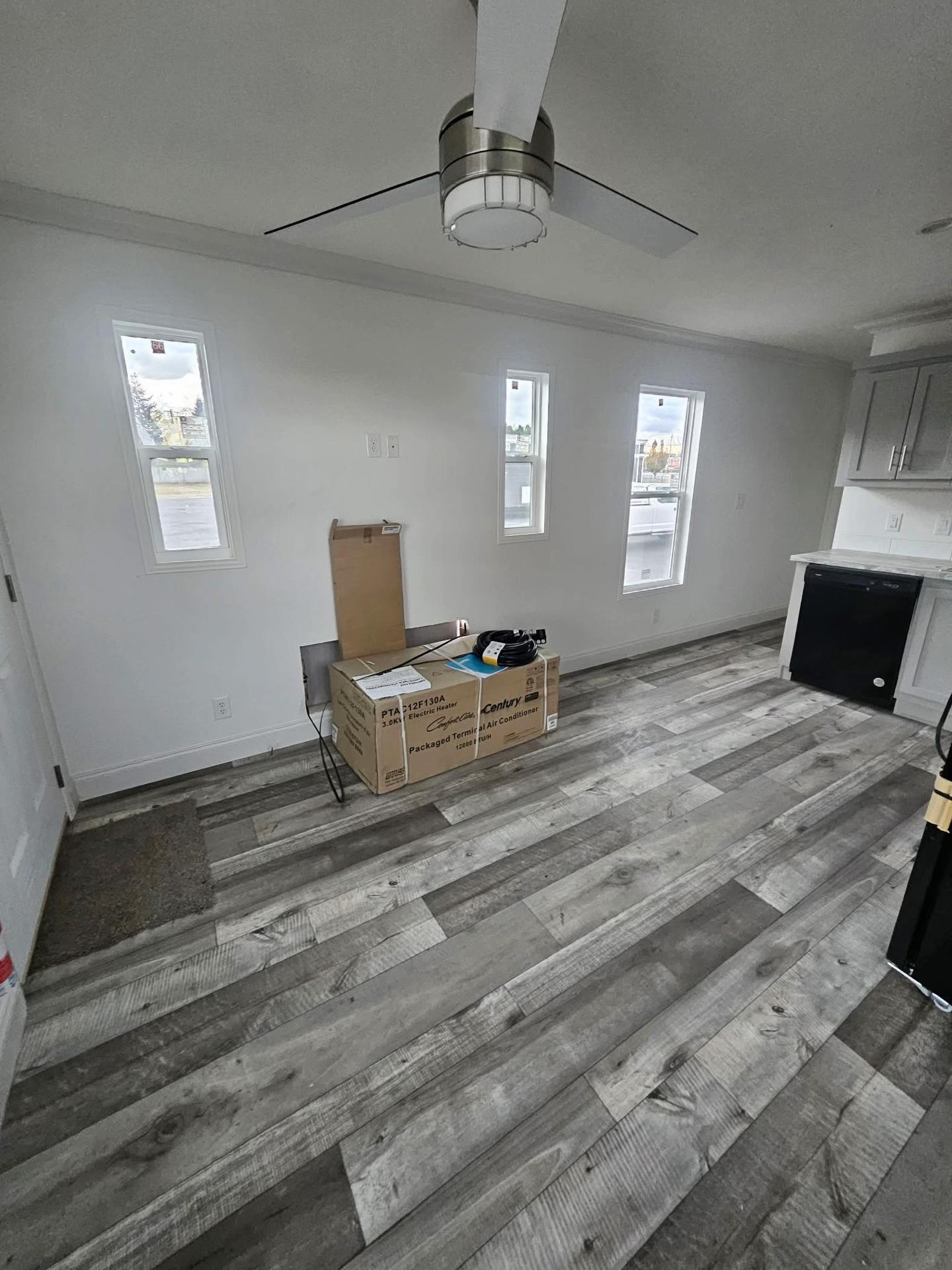 Modern room with grey wood flooring, a ceiling fan, and three tall windows. Cardboard boxes are stacked near the wall, suggesting a move-in.