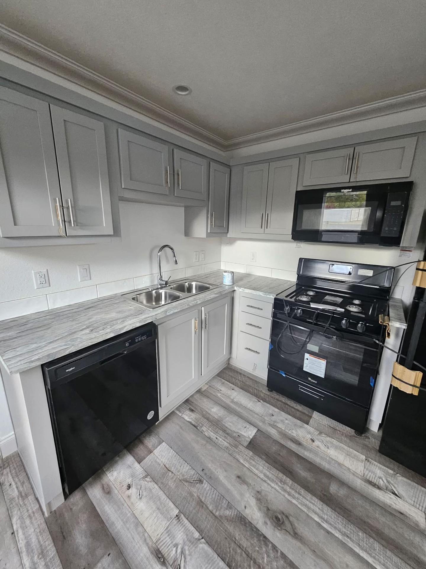 Compact kitchen with gray cabinets, marble countertops, and wood floors. Features a double sink, black stove, microwave, and dishwasher. Modern and neat.