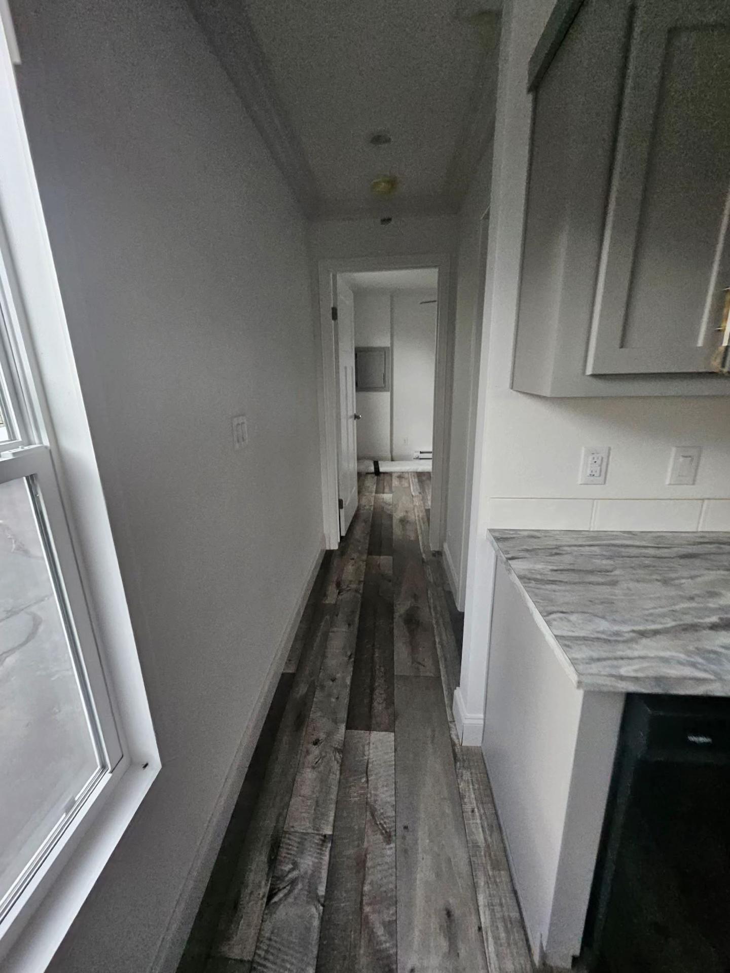 Narrow hallway with wooden flooring, white walls, a marble countertop on the right, and a window on the left. The door at the end leads to another room.