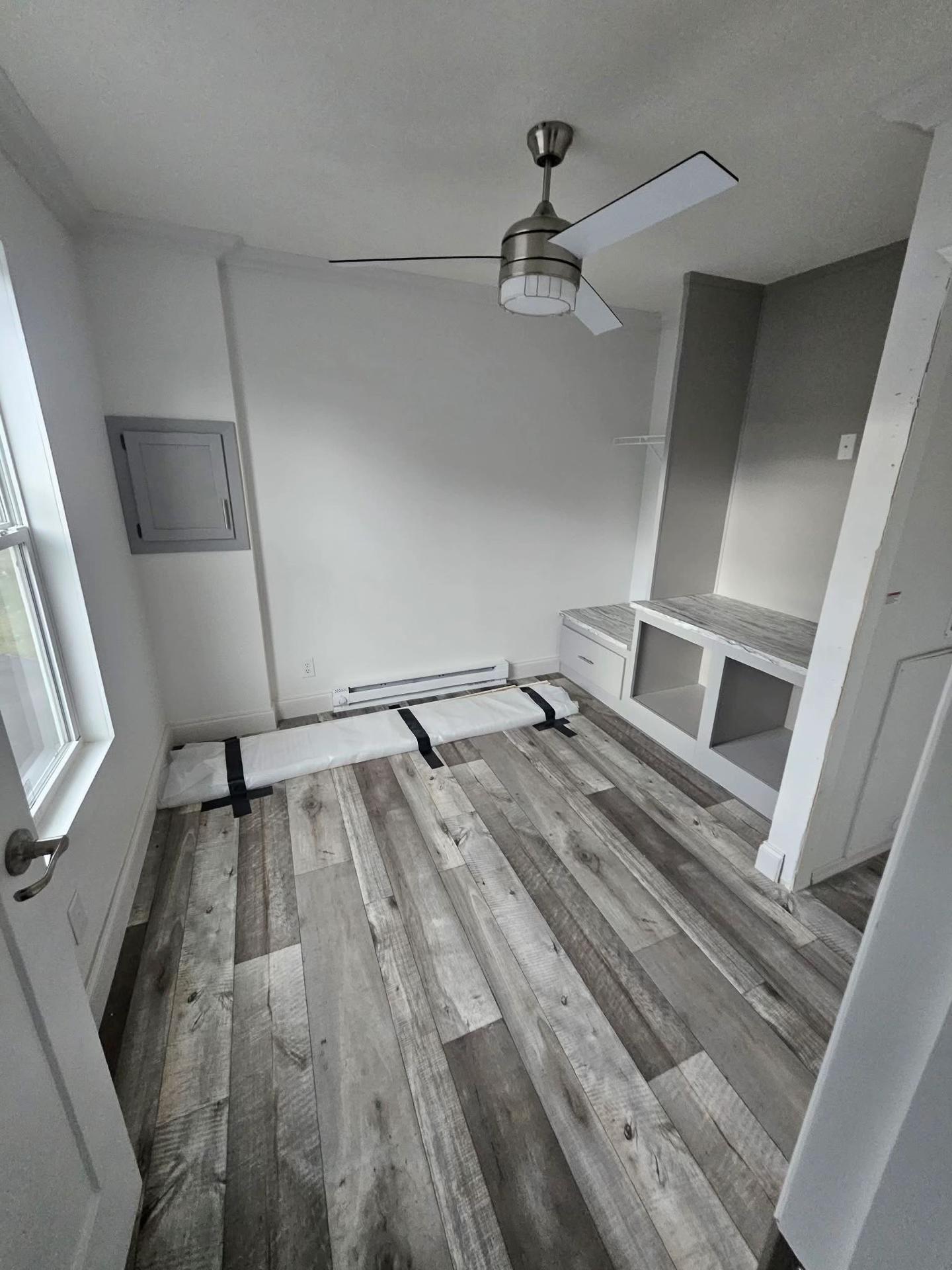 A small room with light gray walls and wood-patterned vinyl flooring. It features a modern ceiling fan, a wall heater, and a built-in desk with shelves. The atmosphere is minimal and clean.