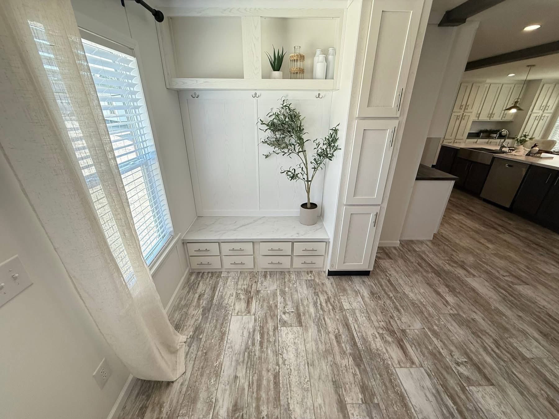 Bright, modern entryway with white cabinetry, hooks, and decor. Features a potted plant, wooden flooring, and leads into a kitchen. Airy and inviting ambiance.