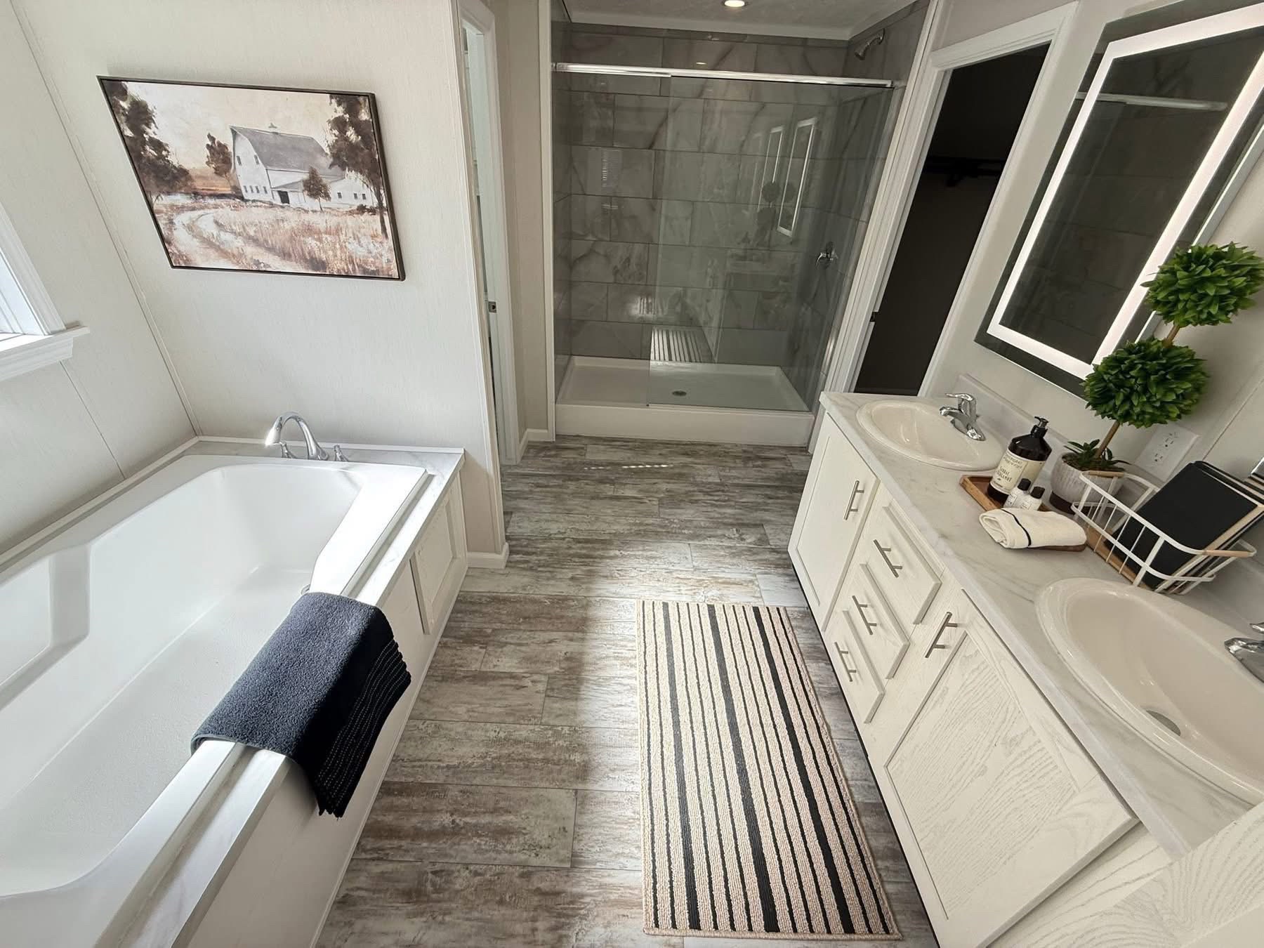 Bright bathroom with a white bathtub, striped rug, and dual sinks. Modern shower, farmhouse painting, plants, and clean, neutral tones create a serene atmosphere.