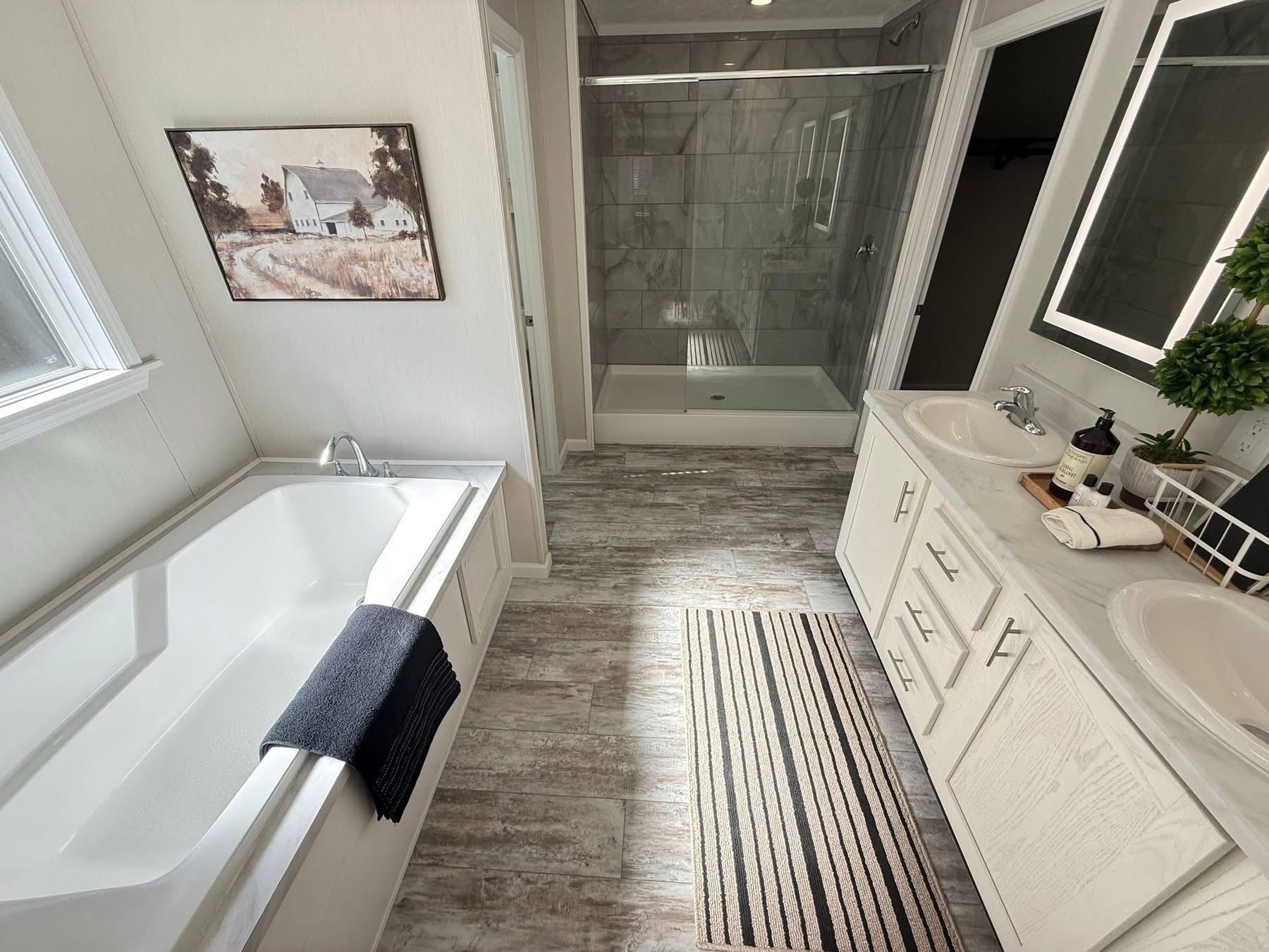 Bright bathroom with a large white tub, black towel, and rustic landscape art on the wall. Double sink vanity with plants and a striped rug are visible.