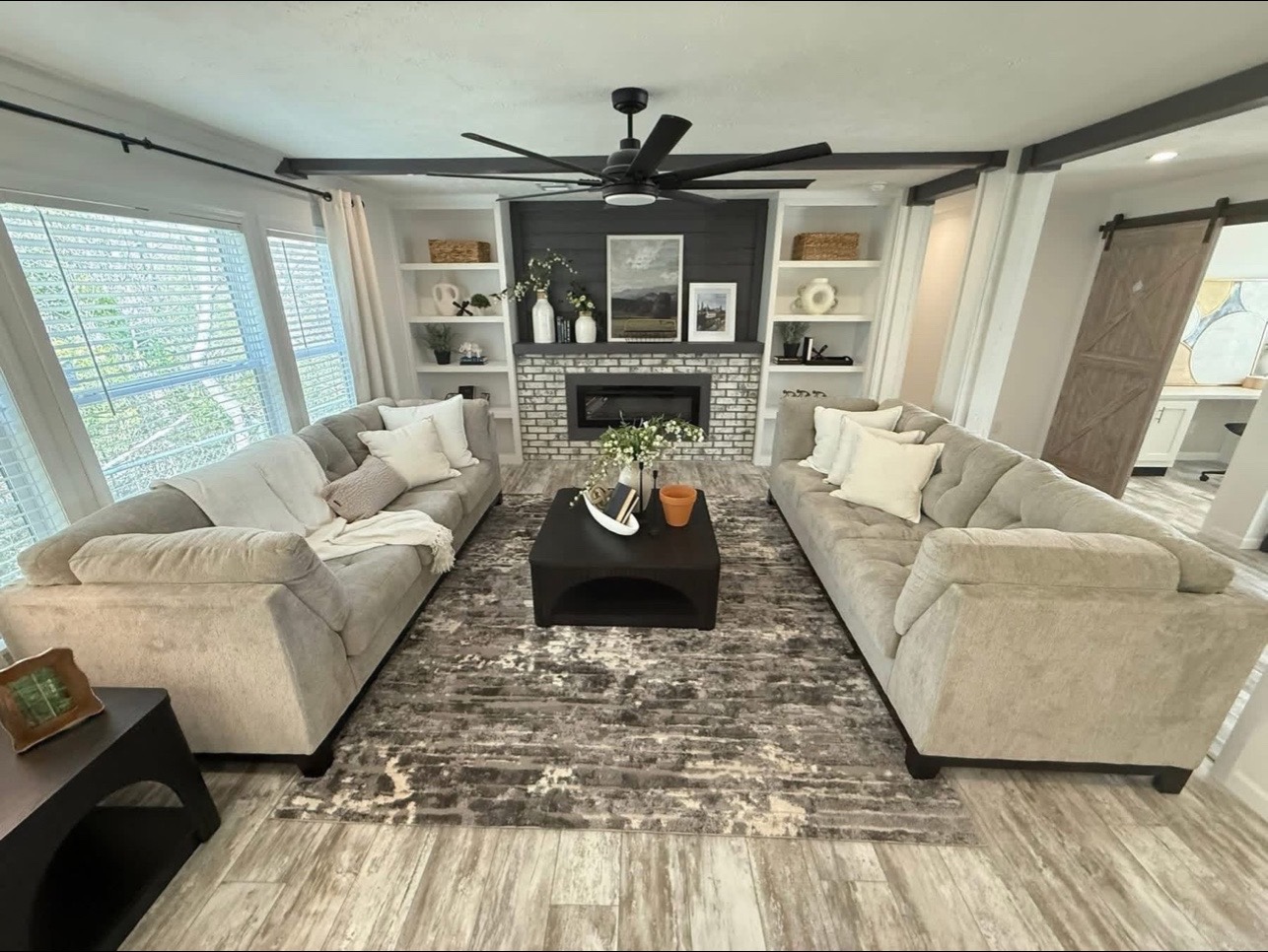 Cozy living room with two beige sofas facing a dark coffee table on a patterned rug. A fireplace with shelves holds decor. Large windows bring light.
