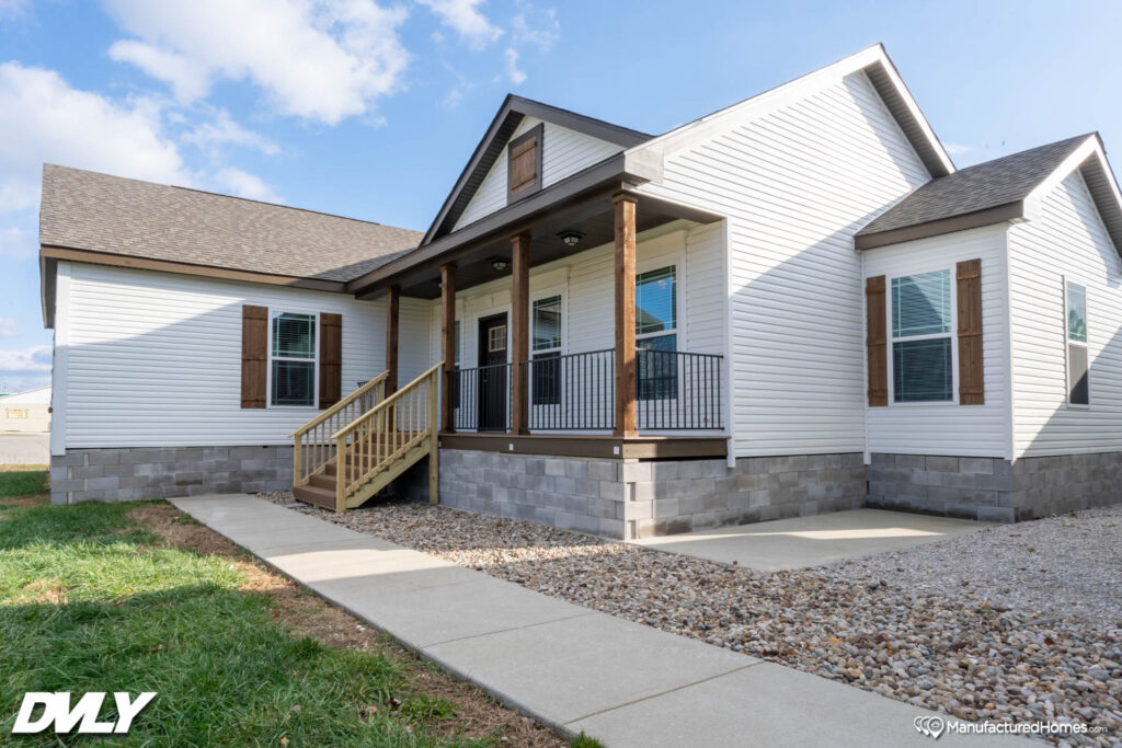 A single-story manufactured home with white siding, wooden shutters, and a small front porch. It has a stone base and a concrete walkway. The sky is clear with a few clouds.