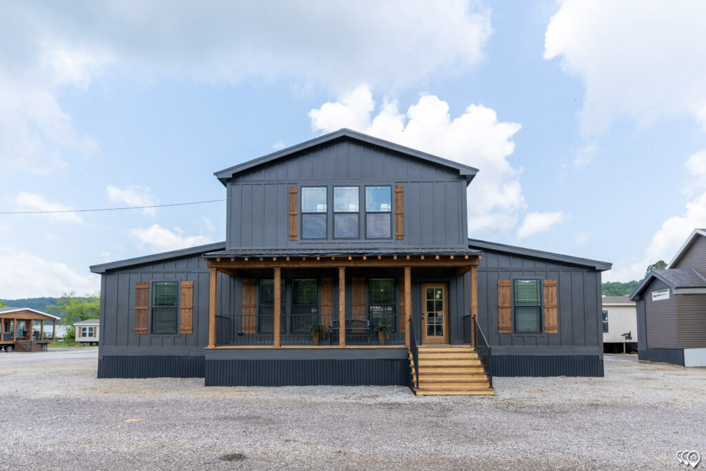 A modern dark gray barn-style house with wooden shutters and a porch featuring steps and railing. Bright sky with scattered clouds in the background.