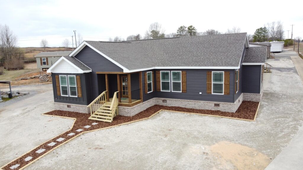A modern, single-story modular home with dark siding, large windows with shutters, front steps, and a neat yard. Overcast sky and barren trees.