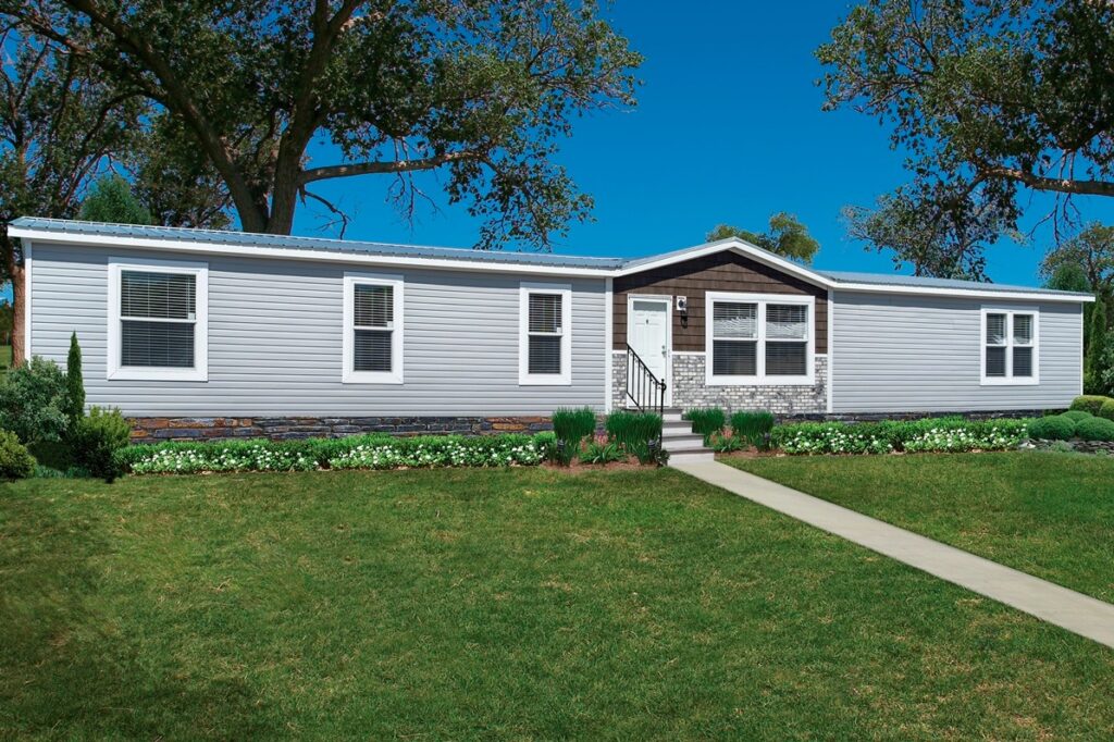 A single-story, white manufactured home with a brown and stone entrance sits amid a well-manicured lawn. A concrete path leads to the front steps, flanked by trees and greenery under a clear blue sky.