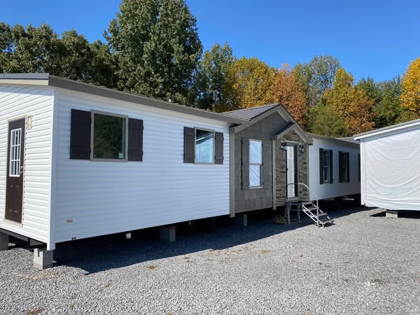 White mobile homes with dark shutters sit on a gravel lot, surrounded by trees with autumn foliage under a clear blue sky. The scene is peaceful and inviting.