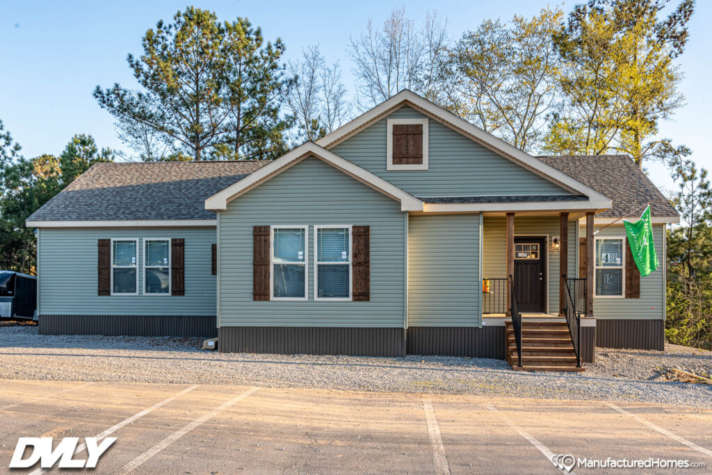 A modern manufactured home with blue-gray siding, brown shutters, and a small porch. It sits on gravel with trees in the background, conveying a serene suburban feel.
