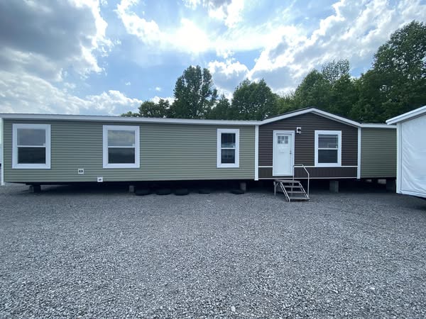 A green and brown mobile home with white trim sits on a gravel lot under a partly cloudy sky. Trees are visible in the background, creating a calm atmosphere.