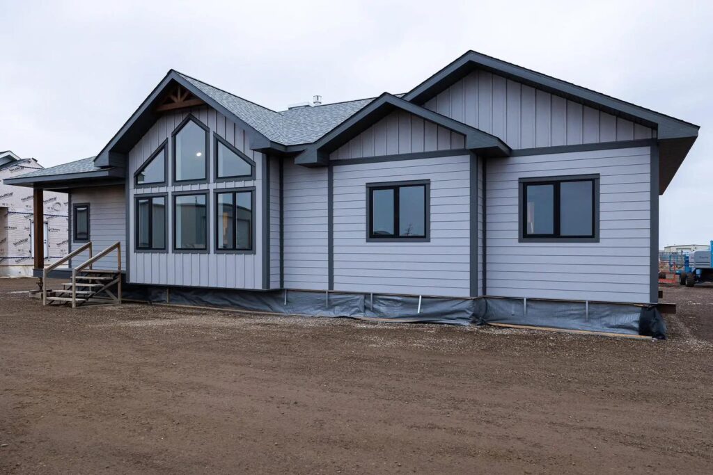 Gray single-story house with large windows and dark trim, set on a dirt lot under an overcast sky. Steps lead to the front door, creating a simple, calm atmosphere.