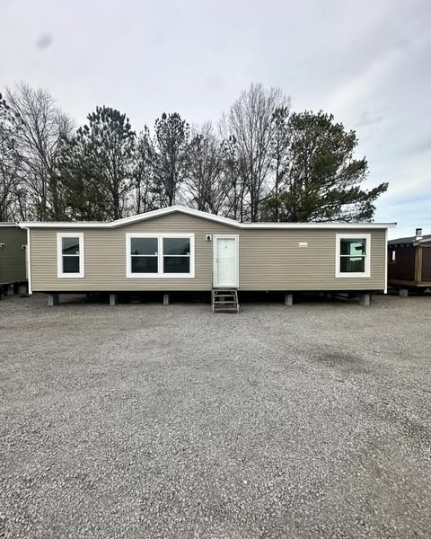 A beige mobile home with white trim, set on gravel, surrounded by leafless trees under a cloudy sky, exudes a quiet and simplistic tone.