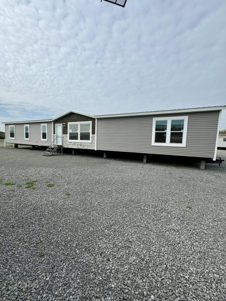 A long, single-story mobile home with gray siding and large windows sits on a gravel lot. The sky above is cloudy, creating a calm atmosphere.