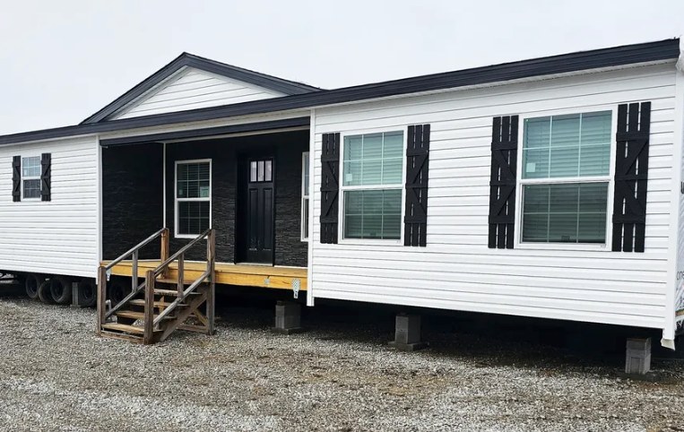 A white manufactured home with a dark roof and shutters sits elevated on blocks. It features large windows and a wooden porch with stairs on gravel.