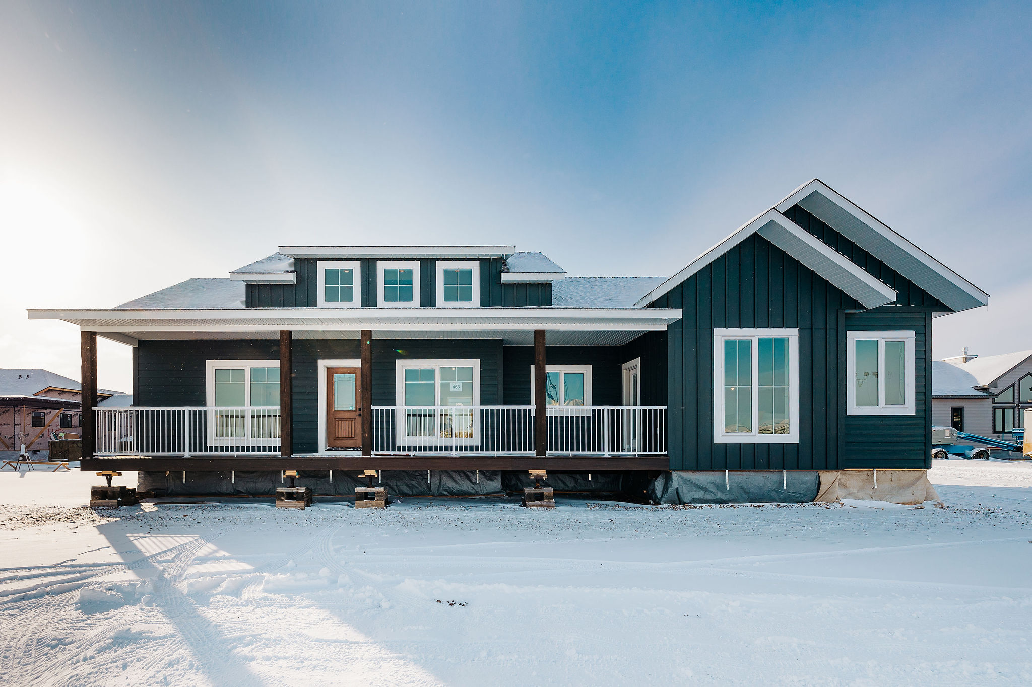 A dark blue house with a large porch and white trim stands in a snowy landscape under a clear blue sky, evoking a serene wintery feel.