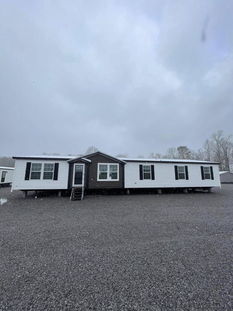 Single-story white mobile home with dark trim and multiple windows, set on a gravel lot under an overcast sky. Sparse trees are visible in the background.