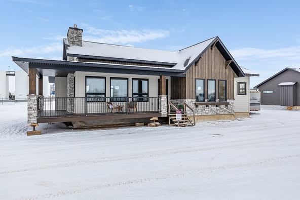 A modern house with large windows and a wraparound porch, set in a snowy landscape. The exterior features stone accents and wood paneling under a clear blue sky.