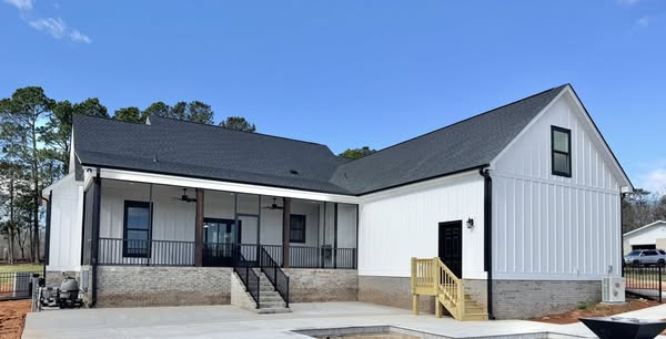 Modern farmhouse-style home with white siding and dark roof. A brick base and black accents add contrast. A porch with stairs leads to the entrance. Bright, clear sky above.