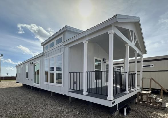 Modern tiny house with white siding, large windows, and a small covered porch under a bright blue sky. Sun shines behind the roof, casting gentle shadows.
