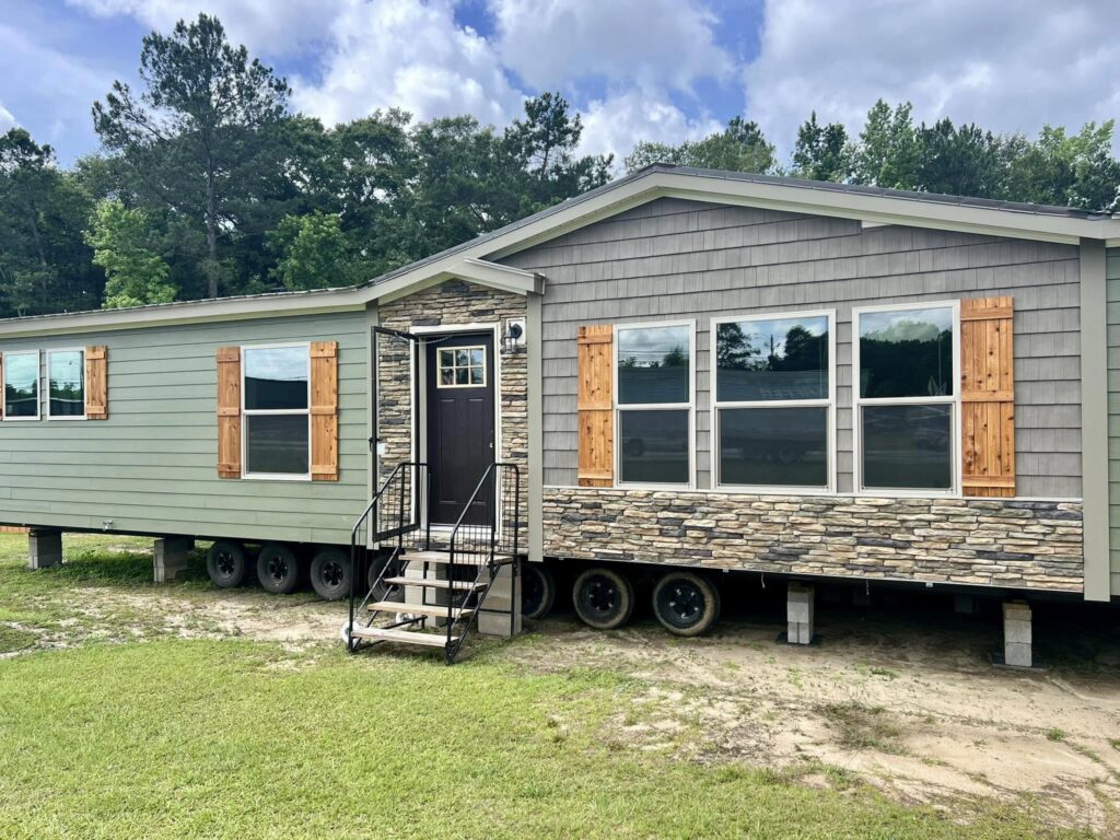A mobile home with stone accents and wooden shutters on windows. It sits on wheels with a small black staircase leading to the entrance, set against a backdrop of trees under a cloudy sky.