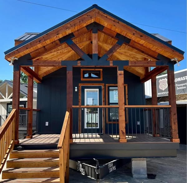 Small wooden cabin with a modern design. Features a triangular roof, dark exterior, prominent wooden beams, and a welcoming front porch. Bright, clear day.