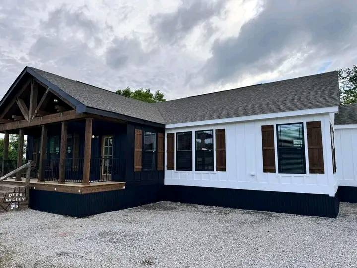 Modern house with contrasting black and white siding and wooden shutters. Covered porch with wooden beams. Gravel foreground under a cloudy sky.